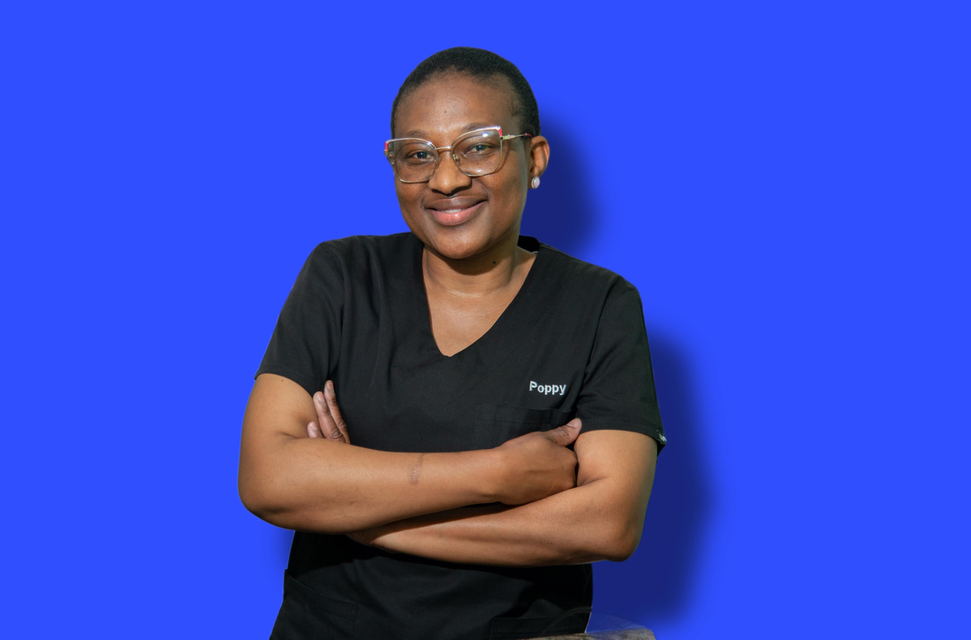 Poppy Koena, the practice manager at Dr T Matseke Practice, smiling with arms crossed in black scrubs against a blue background